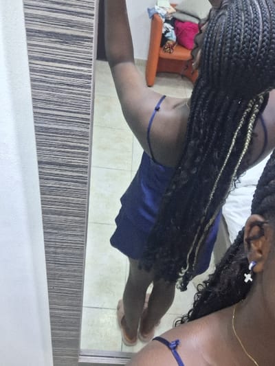 This image depicts a domestic interior in the city of Close, Nigeria. The primary subject is an individual, seen from behind and partially reflected in a mirror, standing on a light-colored tiled floor. The individual has dark skin and long, dark braided hair, some of which feature lighter, possibly golden, interwoven strands. They are wearing a dark blue, thin-strapped sleeveless garment, likely satin or silk, and light-colored slip-on sandals. A gold-colored chain necklace is visible, along with a cross-shaped earring and another small earring on the visible ear. The individual's left arm is raised. The mirror, framed with a horizontal striped pattern, reflects the subject and a cluttered background. This background includes an orange armchair with various folded or discarded clothing items (including blue, pink, and red fabrics), a plain white wall, and a glimpse of what appears to be a white sheet or bedding on the far right. The scene suggests a casual, indoor setting, possibly a bedroom or dressing area.