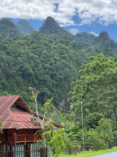 The image depicts a rural landscape with a focus on a traditional wooden house nestled at the foot of a lush, green mountainside. The house features a red corrugated metal roof and wooden railings on a raised porch. A fence surrounds the property. The mountains are densely covered with trees and vegetation, with rocky outcrops visible in places. The sky is partly cloudy with blue patches. A paved road runs along the bottom right of the frame, suggesting a well-traveled path. The scene appears to be located in the city of Thuận Hòa Commune, Vietnam, given the style of the architecture and the tropical vegetation. The overall impression is one of tranquility and natural beauty.