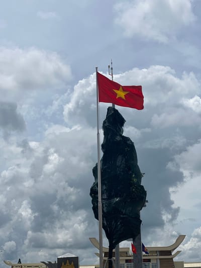 An outdoor, low-angle, full shot captures the Vietnam flag waving on a flagpole in front of a large, dark, textured monument covered in dark green netting. The monument's irregular shape suggests it may be a rock formation or sculpture. The flag, red with a yellow star, is positioned in the upper portion of the frame, against a cloudy, overcast sky. Below the monument, the facade of a building is visible with signs in Vietnamese. To the left of the monument, a white, curved architectural element is seen. The location is the Ben Cau border crossing in Ben Cau, Vietnam.