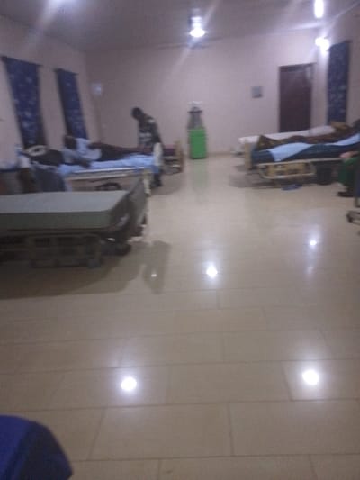 The image depicts an interior scene, identified as a medical ward or similar facility in Nibo, Nigeria. The room is illuminated by multiple overhead lights, which cast bright reflections on the light-colored, tiled floor. The walls are plain and light-toned.
On the left side of the room, two beds are visible. The closest bed in the foreground is empty, featuring a grey mattress on a metal frame. Further back, a second bed is occupied by a person lying down, covered by a blue sheet. Standing beside this bed, facing the occupant, is an individual wearing a striped top, appearing to attend to the person in the bed. Two vertical blue curtains hang against the left wall.
In the mid-ground on the right side of the room, two additional hospital beds are present. One bed is occupied by a person covered with a brown blanket and blue sheet. A dark brown door is visible on the far right wall. A green object, resembling a water dispenser or a medical cart, stands in the background toward the center-right.
A segment of dark blue fabric is visible in the lower-left foreground. No direct interactions beyond the individual standing by the bed on the left are distinctly observable.