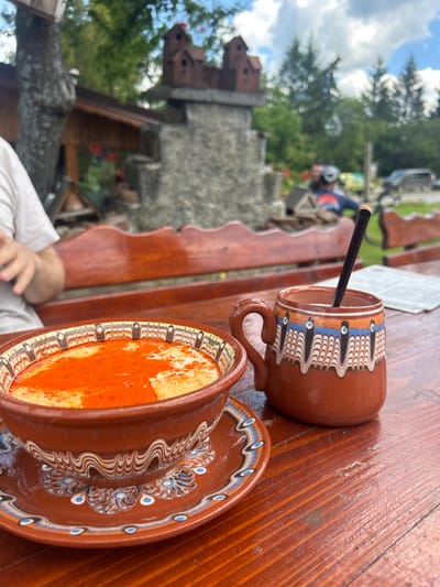 The scene captures a moment at the Tavern "Prista" in Meshtitsa, Bulgaria. A wooden table prominently features two ceramic cups and saucers with a distinct brown hue and intricate white and blue designs. One cup contains a dark straw, while the other holds a beverage with a vibrant orange hue, possibly topped with a powder. In the background, blurred figures of people are visible, suggesting an outdoor setting. The architecture in the background features a stone structure with a castle-like design, adding to the location's atmosphere. Trees and greenery are present, contributing to the natural surroundings of the tavern. A glimpse of a cloudy sky is captured above the trees.
