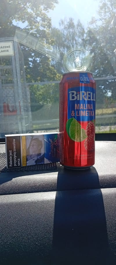 The image displays the interior of a vehicle, specifically the dashboard, viewed from within, with the exterior visible through the front windshield. The scene is illuminated by strong sunlight, casting defined shadows.

On the dark, textured dashboard, two primary objects are present:
1.  **A Beverage Can:** A red and blue aluminum can, open with the tab pushed inwards. The brand "BiRELL" is prominently displayed, along with "MALINA & LIMETKA" (Raspberry & Lime) indicating the flavor. The can is marked "ALK. 0,0% OBJ." (0.0% alcohol by volume) and "S VITAMINY B6 & C" (with vitamins B6 & C) and "BEZ KONZERVANTU" (without preservatives). Manufacturer information for "Česká republika" and "Slovenská republika" is partially visible, indicating a Czech origin.
2.  **A Pack of Cigarettes:** A rectangular white, blue, and brown cigarette pack, positioned horizontally to the left of the can. The brand "Davidoff 100's EVOLVED BLUE" is visible. The pack displays two health warnings in Polish: "Palenie jest przyczyną zawałów serca" (Smoking causes heart attacks) and "Dowiedz się jak rzucić palenie: 801 108 108 www.jakrzucicpalenie.pl" (Learn how to quit smoking: 801 108 108 www.howtocuitsmoking.pl). An image depicting a deceased man, a standard graphic health warning, is also visible on the pack. The cigarette pack rests on a black, textured rubber mat or pad.

The exterior environment, viewed through the windshield, shows a bright blue sky with green, deciduous trees. Sunlight creates reflections and glare on the windshield surface. A prominent reflection of a sticker or label, including a barcode and mirrored text, is visible on the upper left-middle portion of the windshield. On the far left, a white rectangular sign with black text, "PRÁZDNÉ LAHVE" (Empty Bottles), is visible, supported by a metal frame, indicating a bottle return point. The vehicle is situated in Karviná, Czechia.