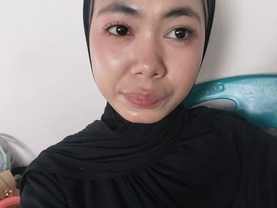 A close-up selfie shows a young woman wearing a black hijab and black garment. She has fair skin with blush on her cheeks and noticeable mascara on her eyelashes. Her lips are slightly parted, and she is looking directly at the camera. The background is a plain white wall, with a blue plastic chair visible on the right side of the frame. The lighting suggests an indoor setting. There is no information available to determine the location in the city of Jeppee, Indonesia.