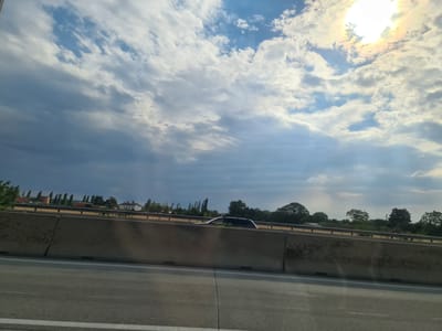 The image captures a view from a vehicle traveling on a highway, likely in or near Wiener Neudorf, Austria, given the provided context. The foreground shows a section of the highway asphalt, marked with white lines. A concrete barrier separates the lane from the opposing direction of traffic, which is visible in the middle ground. Beyond the barrier, a dark-colored car is observed driving in the opposite lane. The background features a landscape with trees and buildings, including some industrial structures and houses, suggesting a semi-urban or rural environment. The sky above is filled with dramatic clouds, with the sun partially obscured, creating a bright halo effect and casting rays of light. The overall scene depicts a typical highway travel view with a dynamic sky.