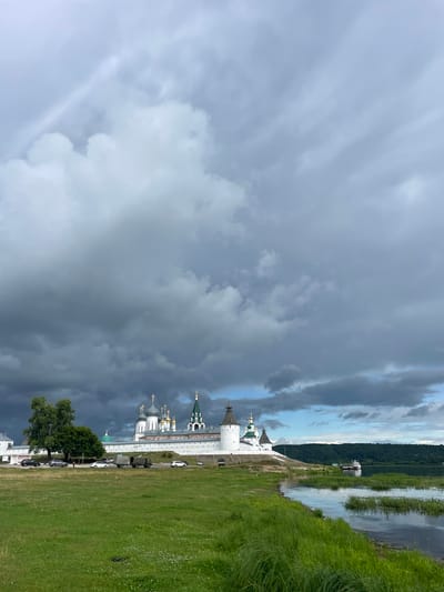 The image shows a view of Makar'evo, Russia. In the foreground, a grassy field with some scattered white flowers slopes down to the edge of a calm body of water. The water reflects the sky and the vegetation along its banks. In the middle ground, a white monastery with numerous domes and towers stands on a slightly elevated embankment. The monastery has white walls and distinctive green and gold domes, some topped with crosses. Several cars and a military truck are parked along a road in front of the monastery. A large, lush green tree stands to the left of the monastery. In the background, a densely forested hill rises on the opposite side of the water. The sky above is filled with dramatic, dark gray storm clouds, with patches of lighter sky visible in the distance. A few boats are docked near the shore on the far side of the water. The scene depicts a historical and religious site under a brooding sky.