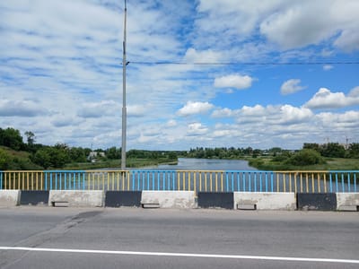 The image depicts a view from a bridge overlooking a river in Stryzhavka, Ukraine. The bridge has a railing composed of alternating blue and yellow vertical bars, with a weathered concrete barrier beneath it. The road surface is asphalt, marked with a white line. Beyond the bridge, a calm river flows, bordered by lush green trees and vegetation. In the distance, there are a few buildings and what appears to be construction cranes. The sky is filled with scattered white clouds against a bright blue backdrop. A solitary utility pole stands prominently on the left side of the bridge. The scene appears to be a daytime shot during fair weather.
