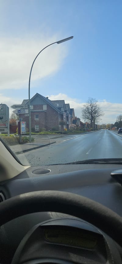 The image captures a view from inside a vehicle, likely a car, driving on a road in Rietberg, Germany. A steering wheel is visible on the right side of the frame. The road is wet, reflecting the sky, suggesting recent rain or wet conditions. On the left side of the road, there are residential buildings with gabled roofs constructed of brick. A street lamp with a curved arm stands near a signpost with multiple signs. The signpost contains direction indicators. The sky is visible with some clouds. Buildings and trees are in the background.