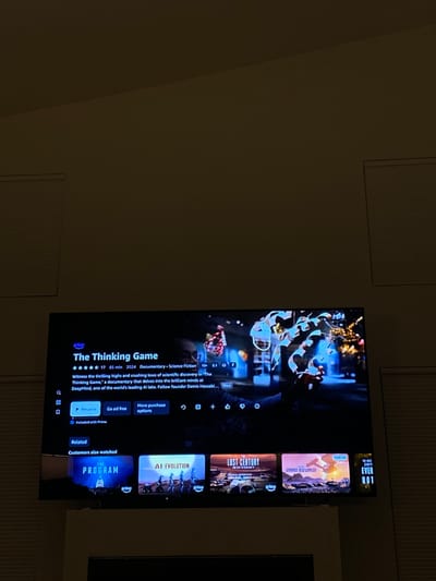 The image depicts an indoor scene within a dimly lit room located in Ridgefield, United States. A large, black flat-screen television is centrally positioned, displaying the Amazon Prime Video interface. The screen prominently features information for a documentary titled "The Thinking Game," indicated as released in 2024, 85 minutes in duration, with a 5-star rating (from 17 reviews), and categorized as Documentary and Science Fiction, rated 13+. The synopsis visible mentions DeepMind and its founder Demis Hassabis. The background image associated with "The Thinking Game" shows a person in silhouette interacting with a glowing, intricate blue and white abstract structure. Below the primary content, a "Related" section displays thumbnails for additional titles, including "THE PROGRAM," "AI EVOLUTION," "THE LOST CENTURY," and "STARRED: FELL WORLDS." The television is situated above a wide, light-colored structure, likely a fireplace mantle or media console. The walls are light-toned. Two windows, one on the left and one on the right, are visible in the upper corners, each covered by closed horizontal blinds. No human subjects are present in the frame, and no active interactions are occurring.