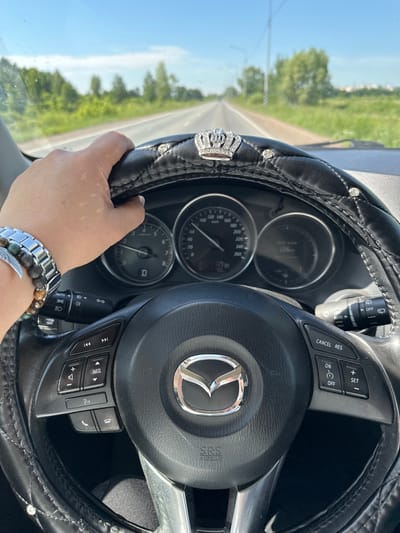 A first-person perspective shot shows a hand with a silver and beaded bracelet on the wrist, gripping a black steering wheel adorned with a jeweled crown emblem and diamond-like accents. The steering wheel has various buttons for media control and cruise control. The dashboard displays gauges with a needle indicating speed. The background is a blurred view of a road stretching into the distance, bordered by green trees and a bright blue sky. The scene suggests the person is driving on a highway or a rural road. The location is identified as Milovka, Russia.