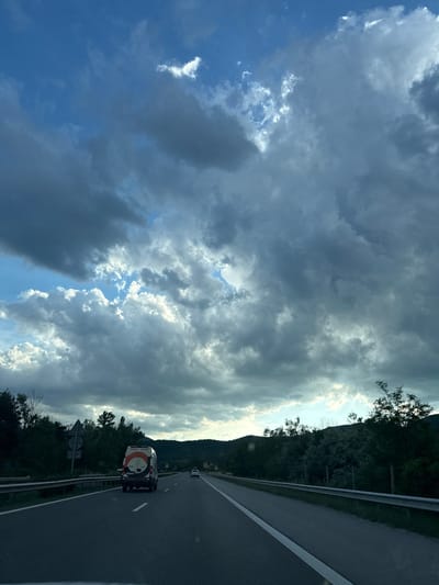 The image depicts a highway scene in the vicinity of Gorno Varshilo, Bulgaria. The sky is filled with dramatic, dark clouds interspersed with patches of bright blue sky and illuminated clouds, suggesting a dynamic weather condition. A multi-lane asphalt highway stretches into the distance, with white lane markings visible. In the foreground, a white van with a distinctive orange and black circular logo on its rear is driving in the right lane. Further ahead, a white passenger car is visible in the distance. The highway is bordered by a metal guardrail and lush green trees and vegetation lining the sides, with rolling hills in the background. The overall impression is one of travel through a scenic, somewhat overcast landscape.