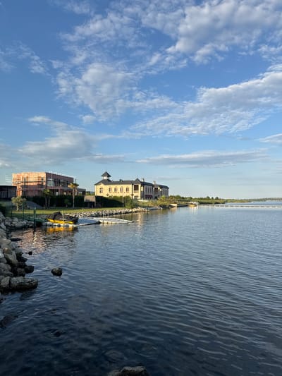 This is an exterior, eye-level, landscape, vertical shot of a waterfront property in Kazashko, Bulgaria. The scene depicts a calm, rippling body of water in the foreground, with a rocky shore to the left. Several boats are docked on the water. In the middle ground, there are two buildings: one is a large, ornate, yellow stone structure with a dark, pitched roof and multiple windows. The other is a modern building under construction, featuring exposed brick and scaffolding. Palm trees and manicured green lawns are visible between the buildings and the water. The background shows a distant shoreline with more buildings and vegetation, all under a bright blue sky with scattered white clouds.