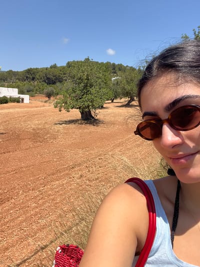 The image shows a close-up of a young woman wearing sunglasses and a light blue tank top, with a red bag strap visible. She is outdoors, with a dry, reddish-brown field stretching out behind her. In the background, there are trees and a white building with a flat roof. The sky is clear and blue. The scene appears to be in a rural or agricultural area, possibly in or around Santa Eulària des Riu, Spain, given the characteristic dry, Mediterranean landscape.
