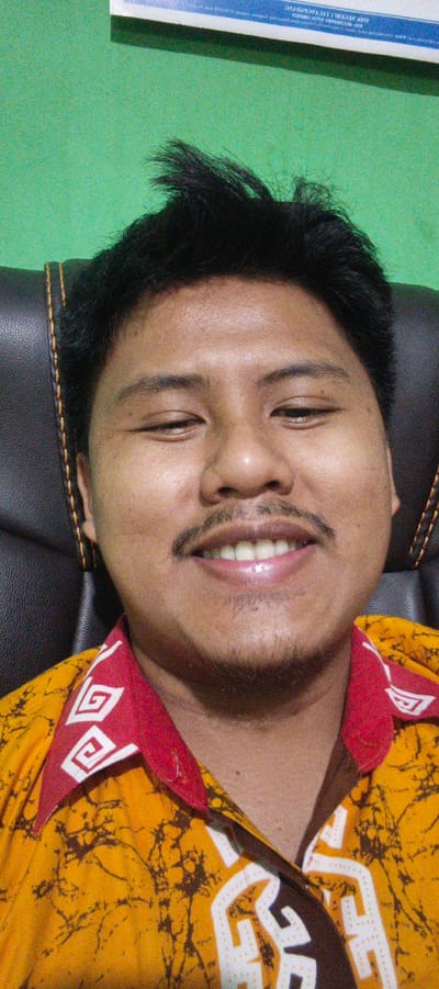 A close-up selfie of a man in Sukabumi, Indonesia, shows him seated in a black leather chair against a bright green background. The man has dark hair, a mustache, and is wearing a yellow shirt with a red collar adorned with white geometric patterns. He is looking directly at the camera with a slight smile, his eyes partially closed. The lighting is somewhat harsh, creating visible skin texture and highlights. A portion of a white sign with text in a foreign language is visible at the top of the frame.