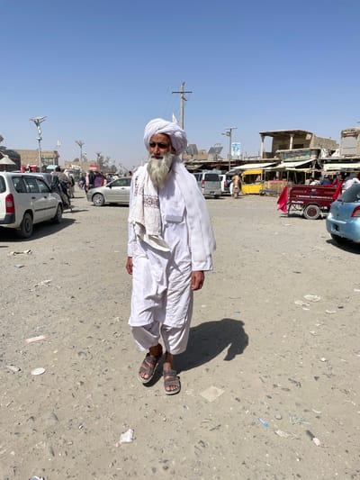 An elderly man with a long white beard and dark sunglasses walks forward across a dusty, unpaved street in Spin Boldak, Afghanistan. He is dressed in traditional white clothing, including a white turban, a white tunic, baggy white trousers, and a white shawl draped over his left shoulder, revealing embroidered patterns on a secondary piece of cloth near his chest. He wears dark-colored open-toed sandals. The ground beneath him is light brown, strewn with small pieces of trash and debris, and a distinct shadow is cast to his right side.
In the background, a bustling market scene unfolds under a clear, light blue sky. Various vehicles are present, including a white compact SUV on the left, a silver sedan further back, and a light blue hatchback on the right. A red motorized three-wheeled cargo vehicle, labeled "BERANG MOTORCYCLE," is parked near makeshift market stalls. These low-rise, weathered buildings feature corrugated metal roofs and colorful awnings in yellow, blue, and red. Numerous other individuals are visible in the middle distance, some standing, some appearing to be engaged in commerce or transit. Utility poles with wires and modern-looking streetlights, some equipped with solar panels, punctuate the scene.