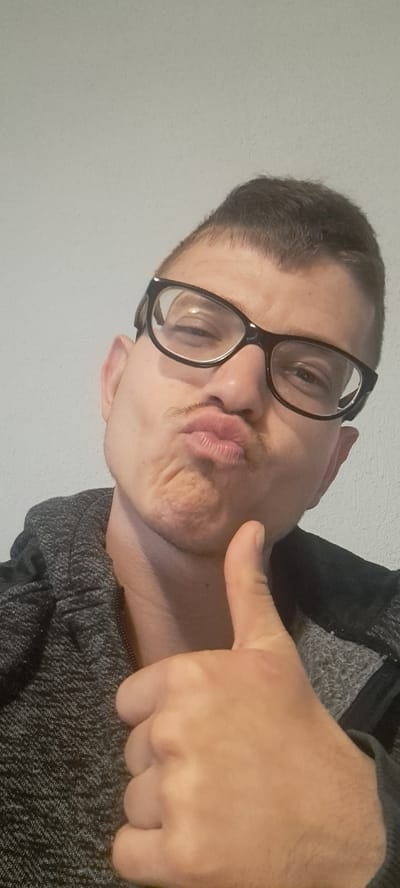 A man with short, dark hair and wearing black-rimmed glasses is positioned at an angle, facing the camera with puckered lips. He is wearing a gray sweater. His right hand is raised, giving a thumbs-up gesture. The background is a plain, light-colored wall. The location is Mineralni Bani, Bulgaria.