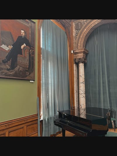 The image depicts an indoor scene, likely a room within a museum or historic building in Saint-Août, France. On the left wall, a large, framed oil painting hangs. The painting portrays a man in formal attire, seated in an ornate chair and holding a document. The man has a dark mustache and is dressed in a black suit with a white shirt and tie. The painting is rendered in a realistic style with rich colors.
In the foreground and to the right, a black grand piano is positioned. The piano is polished and reflects the ambient light. Behind the piano, a dark green curtain hangs, partially obscuring the background. To the right of the piano, a single chair with a dark green seat is visible, suggesting this may be a performance or practice space.
The room features architectural details, including decorative wooden paneling on the lower part of the walls and an arched structure with fluted marble columns supporting it. Above the arch, a circular relief or medallion is visible, adorned with laurel leaves. The overall impression is one of a well-preserved, possibly historical, interior.