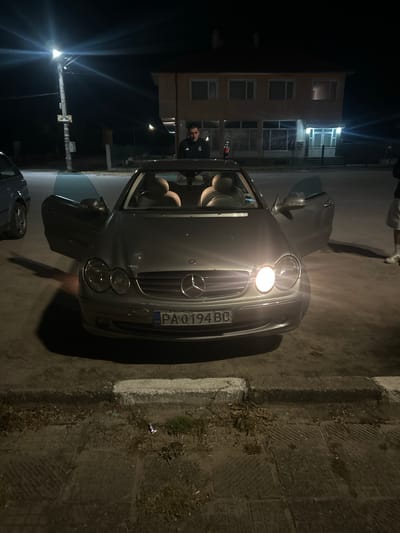 The image depicts a silver Mercedes-Benz parked on a street in Lesovo, Bulgaria, at night. The car has a Bulgarian license plate "PA 0194 BG". The driver's side door is open. A person is standing near the vehicle, and another is partially visible in the background of the building. The street appears to be paved with cobblestones and there is a building illuminated by streetlights.