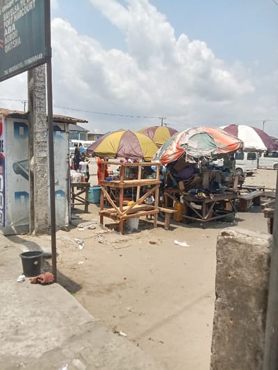 The image shows a street market in Ovwian, Nigeria, located at coordinates 5.496 latitude and 5.797 longitude.  There are several small stalls selling goods under colorful awnings, with some people standing or sitting around them. There is a white van parked in the background. The ground is dusty and sandy, with some trash scattered around. To the left of the frame is a concrete wall with a sign listing several cities in Nigeria. The sign has a black plastic bucket underneath it.  A piece of brown cloth lies in front of the bucket.  The sky is partly cloudy and blue.  A gray concrete post is partially obscuring the view.  The awnings are made of canvas, and some have a  striped pattern.  The stalls are made of wood and have shelves for displaying goods. There is a small blue plastic container in front of one stall.  There is a small wooden table and chair in front of another stall.  The image has a sharp focus on the foreground and a shallow depth of field in the background.