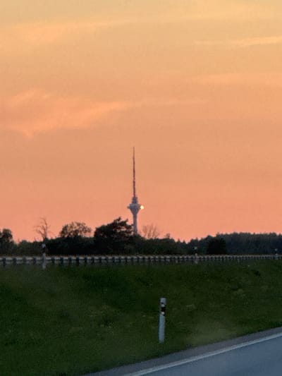 The image shows the Tallinn TV Tower in Jõelähtme, Estonia, at sunset. The sky is a gradient of orange and pink. The TV tower, a prominent structure, is visible in the distance, silhouetted against the sky. In the foreground, there is a grassy area with a fence and a road. The overall scene is serene and picturesque, typical of a rural landscape at dusk.