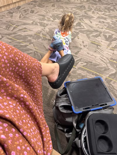 A child with blonde hair is sitting on a carpeted floor, wearing a colorful t-shirt and blue Crocs. Next to the child, a person in a brown floral dress is wearing black slip-on shoes. There is a grey backpack with a blue-bordered tablet on top, and a black travel cooler with two circular compartments. The scene is indoors, likely in a public space like an airport or waiting area, given the carpet and the presence of luggage. The location is Cartersville, United States of America.