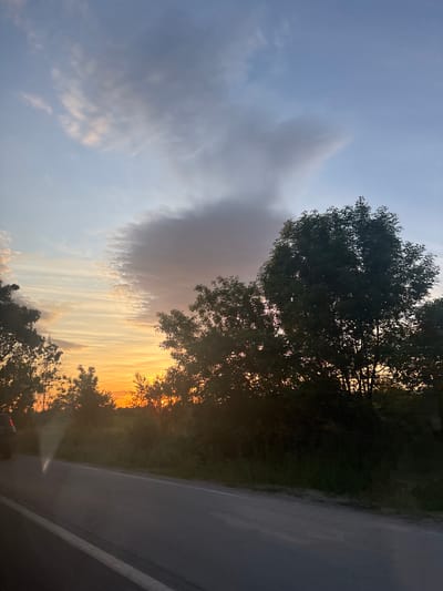 The image shows a view from a moving vehicle, possibly a car, on a road in Stolnik, Bulgaria. The road surface is dark. The right side of the road is visible. In the distance, the sunset creates an orange glow. Trees line the side of the road, silhouetted against the sunset. A large, oddly shaped cloud is on the left side of the image.