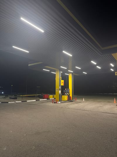 The image shows an outdoor scene at a gas station at night. The overhead canopy of the gas station is illuminated by long, white LED lights. Yellow and black are the dominant colors of the gas station's structure and equipment. Two yellow gas pumps are visible, one on the left with a black screen and red nozzles, and another to the right, also yellow with black accents. Red bollards and yellow barriers are positioned around the pumps. The ground is paved with asphalt in the foreground and interlocking concrete pavers under the canopy. The background is dark, suggesting a rural or suburban setting, with some faint lights and trees visible in the distance. Orange traffic cones are placed on the pavement. The scene is devoid of any people or vehicles, conveying a sense of stillness.