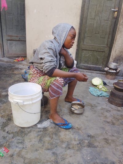 A young woman sits on a stool outside a building. She wears a grey hoodie and colorful patterned trousers. A white bucket sits next to her. In front of her is a silver bowl with food, a cut cabbage, and cooking implements. The woman has her head resting on her hand in a thoughtful pose and wears blue sandals. The area is outdoors with an aged concrete floor.