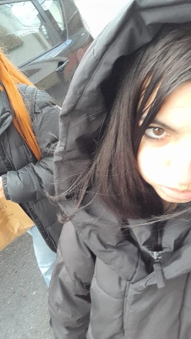 The image captures two individuals outdoors in Meistratzheim, France. In the immediate foreground, a person with long, straight dark brown hair is depicted, wearing a dark grey or black hooded puffer jacket with the zipper partially open. Their right brown eye is clearly visible and directed towards the viewer, while the rest of their face is partially obscured by hair and the jacket's hood. To the left and slightly behind, a second individual with long, orange-red hair is visible, wearing a similar black puffer jacket and light blue trousers. This second person holds a light brown paper bag in their left hand at waist level. In the background, a dark-colored vehicle is partially visible behind the second individual, with its side and a portion of its window reflecting light. The ground surface appears to be paved, resembling asphalt.