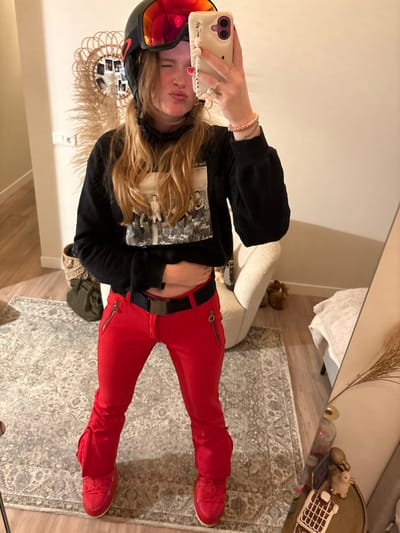 A young woman poses for a mirror selfie. She wears red ski pants with a black belt, a dark sweatshirt, and a ski helmet with tinted goggles. Her long blonde hair falls loosely around her shoulders. She holds her phone up to capture the image. A cream-colored chair is behind her, and a patterned rug is on the floor. Other decorative items can be seen on a nearby table.