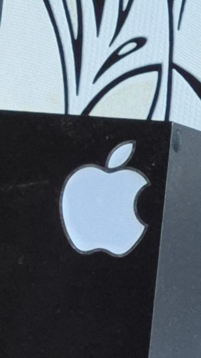 The image displays a close-up of an Apple logo on a black surface, likely a product or packaging. The logo is a light blue silhouette of an apple with a bite taken out of it, outlined in a darker shade. Above the black surface, a white background is visible with some abstract black lines. The overall image is slightly grainy. The location in Neftegorsk, Russia, is not depicted in the image.