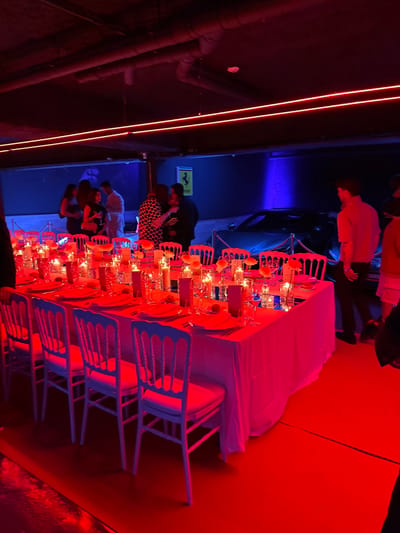 The image shows a long table set for a dinner party in a brightly lit, red-hued room. The table is set with white plates, silverware, and wine glasses. There are white flowers on the table as well as lit candles in various sizes and shapes. The white chairs around the table are mostly empty, as are the seats at the front of the table. Several people are standing off to the side, facing toward the table, and there is a black car in the background. The scene appears to be at an indoor venue at 43.749, 7.429, which corresponds to a location in Beausoleil, France. The bright red lighting makes it difficult to discern further details about the environment.