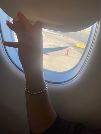 The image depicts a close-up, internal view from an aircraft window seat at Newark Liberty International Airport (EWR), Newark, United States. A human left arm, appearing female, is centrally positioned, with the hand actively manipulating a light-colored, retractable airplane window shade. The arm displays a multi-strand metallic bracelet on the wrist. Outside the oval-shaped window, an airport tarmac is visible under bright daylight, featuring multiple orange traffic cones, yellow ground support equipment, and white ground markings. Shadows are cast across the tarmac, indicating clear weather.