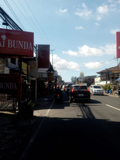 The image displays an urban street in Ban Phonhxavanh, Laos, under a blue sky with scattered white clouds. Bright sunlight casts distinct shadows across the asphalt road, which recedes into the background.

On the left side of the foreground, a large red rectangular sign features white text reading "MAH MAKAN" at the top and "AI BUNDA" below. Further details on this sign include "No. 68 Di depan DST Factory Outlet" and "Telp. (0281) 68484". Below this, a partially obscured red sign also displays "AKAN BUNDA". A dark metal fence and some small green plants are visible in the immediate lower-left foreground. Behind these signs, a continuous row of commercial buildings lines the street, adorned with various other advertising billboards, including yellow and red ones; the word "SEMBIRING" is legible on one of the yellow signs. Numerous overhead utility lines and cables are strung across poles above the street.

Traffic is present on the road, with vehicles moving away from the viewer. A dark-colored SUV is prominently positioned in the central lane. Several other cars are visible behind it. In the left lane, a motorcyclist wearing a red helmet is riding a dark motorcycle. Further down the road on the right, a white sedan is visible among other vehicles. No clear interactions between individuals are discernible.