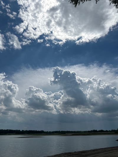 This is a vertical, eye-level, outdoor photograph of a body of water with a treeline in the background and a dramatic sky filled with clouds. The body of water occupies the lower half of the image and appears to be a lake or river, with a sandy shoreline visible in the lower right corner. A dark, dense treeline stretches across the horizon, obscuring any distant landscape.

The sky is the dominant feature of the photograph, with a complex arrangement of clouds. The upper portion of the sky is a deep blue, interrupted by scattered white and grey clouds. Closer to the horizon, towering cumulus clouds are prominent, with some areas appearing bright and illuminated by sunlight, while others are darker and suggest the possibility of rain. The lighting conditions suggest it is daytime, likely mid-afternoon, with the sun partially obscured by the clouds, creating a dynamic contrast.

There is no indication of the location being in the city of Pravda, Russia. The scene appears to be a natural landscape. No people, actions, or interactions are depicted. The image is purely a scenic landscape photograph.