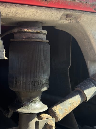 The image shows a close-up of a vehicle's suspension system, specifically an air spring and a shock absorber. The air spring is a black, ribbed bellows-like component, suggesting it is part of an adjustable suspension system. It appears to be mounted to a red vehicle body above and connected to a metal post below. The shock absorber is a rusty cylindrical component, angled diagonally in the frame, with visible markings. The surrounding environment is dark and appears to be the undercarriage of a vehicle. The image was taken on an outdoor surface, likely a road or a driveway, in Zarech'e, Russia. There are no people or any other discernible objects or actions in the image.