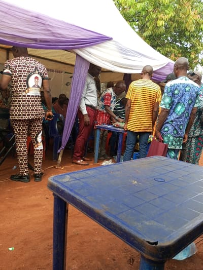 The scene depicts an outdoor gathering in Oko Town, Nigeria. A temporary white fabric tent, draped with purple cloth, provides shade over a group of people. The ground is exposed reddish-brown earth. In the foreground, a worn blue plastic table with a grid-patterned surface and a chipped corner is positioned. Under the tent, several men are congregated. A man, seen from behind on the left, wears a brown, black, and white patterned outfit featuring a large circular portrait of an elderly man in a red cap on the back of his shirt. To his right, a man in a light grey shirt and red trousers stands. Centrally, a man in a red, white, and blue patterned shirt is interacting with items on a smaller blue table. Other individuals include a man in a yellow and black striped t-shirt and another in a blue, green, and black patterned shirt holding a maroon bag. Partial views of other attendees and blue plastic chairs are visible further inside the tent. Green tree foliage extends above the tent structure. A wooden pole, adorned with purple fabric, supports the tent.