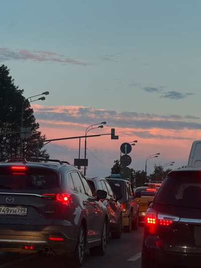 This is a vertical, eye-level, medium shot of traffic congestion in the city of Krasnaya Pakhra, Russia, during a sunset. The sky transitions from a pale blue at the top to a soft pink and orange hue near the horizon, with scattered clouds. Several streetlights with multiple bulbs are visible, some mounted on poles and others on overhead structures. Traffic signals and road signs are also present.
In the foreground, the rear of a dark gray Mazda SUV is prominent, its brake lights illuminated red. The license plate is visible, reading "749YH 799 RUS". To the right of the Mazda, the rear of a dark SUV, possibly a Toyota, is partially visible, with its brake lights also glowing red. In the middle ground, a yellow Yandex taxi with a light bar on its roof is seen, along with other vehicles in traffic. The vehicles are lined up on a paved road. Trees are visible on the left side and in the background, bordering the road. The overall scene suggests a typical evening commute.