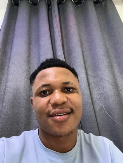 A close-up shot of a smiling man with short black hair. He has a small tattoo above his left eyebrow and is wearing a light blue t-shirt. His skin is clear and light brown. The background features draped gray curtains. The image appears to be taken at an angle, slightly tilted to the left.