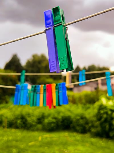 The image shows a clothesline with several clothespins in an outdoor setting. The clothespins are of various colors, including purple, green, blue, red, and yellow. Some are in focus in the foreground, while others are blurred in the background. The clothesline is strung across a blurred green background of foliage, possibly in a garden or park. Above the clothesline, a cloudy sky is visible. The image appears to be taken in Vecumnieki, Latvia, given the context provided. There are no people or animals visible in the image.