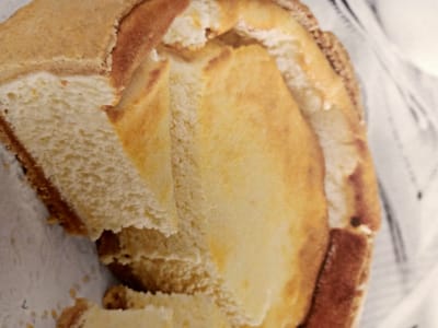 The image shows a close-up, slightly blurry view of a partially eaten sandwich. The sandwich appears to have white bread with a golden-brown crust. The filling is not clearly visible but seems to be a light color, possibly cheese or mayonnaise. The sandwich is lying on a white surface, possibly a paper napkin, with a blurry background that suggests it is in an indoor setting. There are no people or clear indications of a specific location within Rumaithiya, Kuwait. The scene is static and focuses solely on the food item.