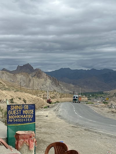 The image depicts a scenic view of a road winding through a mountainous region. In the foreground, a blue sign with yellow lettering announces "SHING-GO GUEST HOUSE BODHKHARBU PH-9419524344." Below the sign, a cylindrical object is covered with a red cloth tied with yellow string. Two plastic chairs are visible in the lower right corner. The road curves to the right, leading into the distance where a green and white truck is traveling.
The landscape is characterized by arid, rocky mountains with sparse vegetation. The sky is overcast with heavy, grey clouds. In the mid-ground, a fence runs along the side of the road, and a few power lines are visible. Further in the distance, a valley with green fields can be seen, nestled between rolling hills and the imposing, dark mountain range that forms the horizon. The overall impression is one of a remote, rugged, and somewhat desolate environment, typical of the high-altitude regions of Ladakh, India, where Bodhkharboo is located.