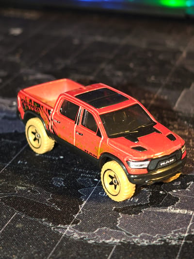 A close-up, eye-level, slightly angled shot focuses on a red toy pickup truck. The truck has black accents, yellow off-road tires, and some gold markings on the side. The truck is positioned on a dark surface that appears to be a map with faint white lines. In the background, out of focus, are blurred colors that suggest a screen or lights. There are no people or actions depicted in the image. The scene is static and does not provide any information about its location in Dörpen, Germany.