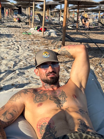 The image shows a man with numerous tattoos lying on a cushion on a beach. He is wearing a baseball cap and sunglasses and has a beard. In the background, there are beach umbrellas, lounge chairs, and other people, indicating a public beach. The scene appears to be a sunny day. The location is identified as Moudros Municipal Unit, Greece.