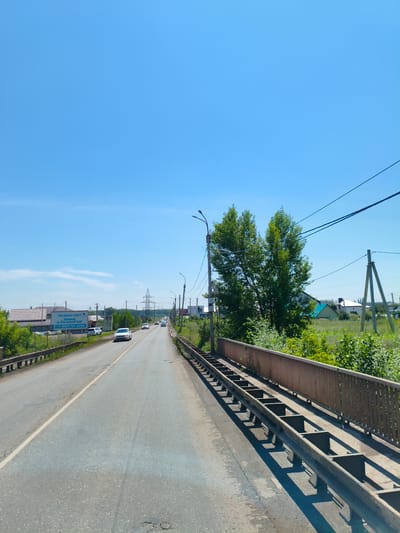 The image shows a bright, sunny day with a clear blue sky over a road in Novaya Otradovka, Russia. The road is asphalt and appears to be a two-lane highway. Several cars are visible on the road, driving in the same direction. On the left side of the road, there are houses with red roofs and lush green trees and vegetation. A large billboard is also visible on the left, with Russian text that appears to be an advertisement. On the right side of the road, there is a metal guardrail and more green trees and bushes. Power lines are strung across the sky, supported by wooden poles. In the distance, there is a large electrical pylon. The overall impression is of a rural or semi-rural setting on a warm day.