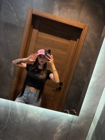 A woman is taking a mirror selfie. She is wearing a pink baseball cap, a black crop top, and blue jeans. She has long dark hair and is wearing makeup. She is holding her phone up to take the picture, and her left hand is raised near her face. She is standing in front of a wooden door and a marble-tiled wall. The mirror is illuminated from above. There is no visible information indicating the location is in Dnp Ryzhiki, Russia.