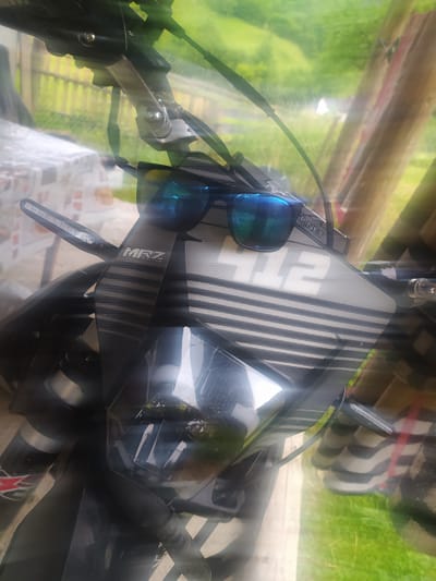 The image shows a close-up, overhead view of a motorcycle handlebar area, likely parked outdoors. The dominant objects are a pair of blue mirrored sunglasses resting on a black and white patterned cover, possibly a seat or part of a fairing. The cover has vertical stripes and a textured surface. To the right of the sunglasses, a smartphone with a clear protective case is visible, its screen reflecting the surroundings. The motorcycle's handlebars and various control cables are also prominent.
Behind the motorcycle, the scene suggests an outdoor setting with greenery, indicating it's likely daytime with bright conditions, possibly sunny due to the reflections and highlights. The background appears blurry, but hints of wooden structures and possibly a fence are discernible, suggesting a rural or semi-rural location.
Visible text includes "2" in large white block letters on the patterned cover, and "MRZ RACING" with smaller text below it, also on the cover. There's also a small text that appears to say "P.NET". The overall impression is of an unattended motorcycle ready for a ride or recently parked. The context of Hoverla, Ukraine, suggests a mountainous or scenic region.