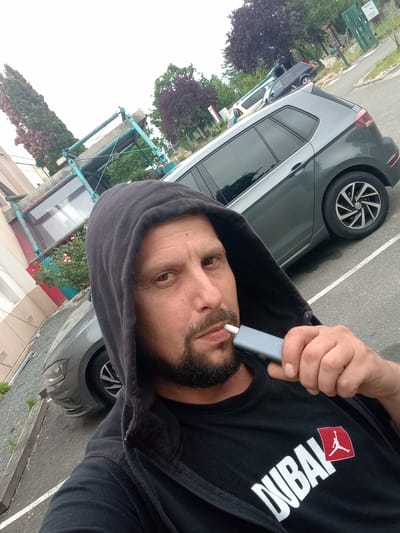 A middle-aged man with a beard, wearing a black hooded sweatshirt and a black t-shirt with "DUBAI" and a Jordan logo on it, is taking a selfie while holding a gray vape pen to his mouth. He is standing in a parking lot. Behind him, there are several parked cars, including a gray Volkswagen. There are trees and a building in the background. According to the metadata, this image was captured in Lormont, France.
