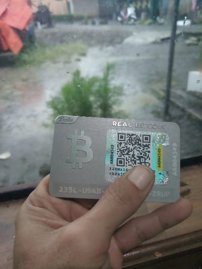 A hand holds a "Real Bitcoin" card. The card is silver and features a QR code under a security sticker. The sticker is partially peeled back. Text on the card includes "AA004549," "12EMz16rbXuJ," and "235L-U9AB." The Bitcoin logo is prominently displayed. The background is blurry, showing a window with an out-of-focus outdoor scene of trees and buildings.