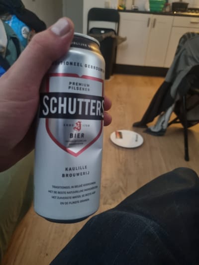 The image captures a close-up of a person's hand holding a can of "SCHUTTERS" beer. The can is primarily silver with red and black labeling. The text on the can indicates it is a "PREMIUM PILSNER" brewed by "KAULILLE BROUWERIJ" and is "TRADITIONEEL IN BELGIË GEBROUWEN" (traditionally brewed in Belgium) with high-quality ingredients.
The background suggests an indoor domestic setting. In the blurred background, a kitchen counter with cabinets and a sink is visible, suggesting the location could be a kitchen. Further in the background, a person wearing dark clothing and possibly grey shoes is seated on a chair. A black trash can is also visible. The flooring appears to be wooden.
The image does not provide any specific details or clues that would place the scene within the city of Haarlem, Netherlands.