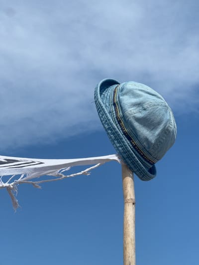 A light blue denim bucket hat is perched on top of a bamboo pole against a bright blue sky with wispy clouds. A tattered white cloth with black zig-zag patterns is attached to the pole, blowing in the wind. The hat has a yellow and dark blue ribbon band around its crown. The scene appears to be outdoors, possibly near a beach or a coastal area.